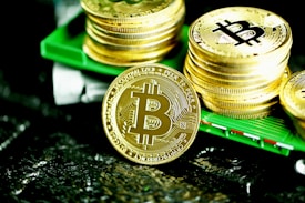A stack of shiny gold coins with the Bitcoin logo printed prominently on each coin. The coins are placed on a green surface, reflecting light, with a prominent Bitcoin coin leaning against the stack.