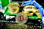 A stack of gold coins featuring the Bitcoin logo is displayed on a miniature green truck. A mechanical blue robotic arm is positioned above, appearing to place or retrieve coins. The scene is brightly illuminated, highlighting the shiny surface of the coins.