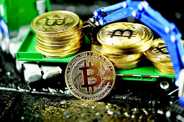 A stack of gold coins featuring the Bitcoin logo is displayed on a miniature green truck. A mechanical blue robotic arm is positioned above, appearing to place or retrieve coins. The scene is brightly illuminated, highlighting the shiny surface of the coins.