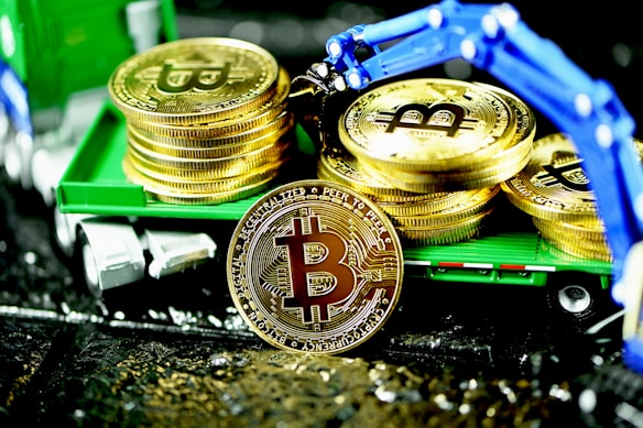 A stack of gold coins featuring the Bitcoin logo is displayed on a miniature green truck. A mechanical blue robotic arm is positioned above, appearing to place or retrieve coins. The scene is brightly illuminated, highlighting the shiny surface of the coins.