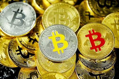 Several gold and silver coins feature the Bitcoin symbol in various colors, including red and yellow. The coins are arranged closely together, creating a dense, overlapping pattern.