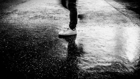 A monochrome image depicting a person walking on a wet, reflective surface. Only the lower part of the person's legs and feet are visible, with the individual wearing dark pants and white sneakers. The surface appears to be slightly textured with patches of light and shadow, emphasizing the water's reflective quality.