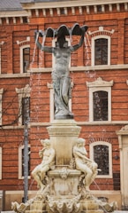 A classical-style fountain featuring a tall sculpture of a standing figure holding a large, shell-like basin above its head. Water cascades from the edges of the basin. Below, two seated figures are sculpted on the fountain's base, surrounded by ornate carvings. The background features a large red-brick building with arched windows and decorative stonework.