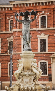 A classical-style fountain featuring a tall sculpture of a standing figure holding a large, shell-like basin above its head. Water cascades from the edges of the basin. Below, two seated figures are sculpted on the fountain's base, surrounded by ornate carvings. The background features a large red-brick building with arched windows and decorative stonework.