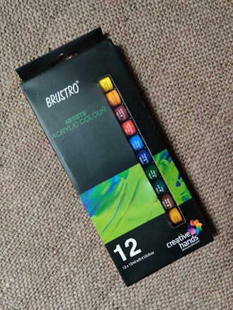 A box of Bruistro artist’s acrylic colours is placed on a textured fabric surface. The packaging is predominantly black with a vibrant, colorful display of different paint tubes along one side, and a section of abstract colorful art near the bottom. The brand name 'CREATIVE HANDS' is printed at the lower right corner. The numbers '12 x 12ml' indicate the quantity and volume of the paint tubes inside.