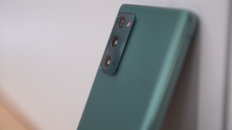A modern smartphone with a sleek green finish and a triple-camera setup on the back. The camera module is prominently featured, and the phone is leaning against a light-colored surface.