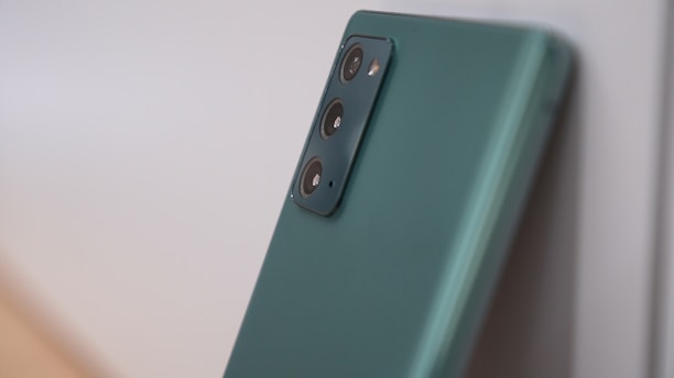A modern smartphone with a sleek green finish and a triple-camera setup on the back. The camera module is prominently featured, and the phone is leaning against a light-colored surface.