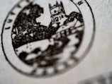 A close-up view of a circular black and white stamp featuring a medieval-style building, possibly a castle, with trees and a house in the background. The details are slightly blurred, emphasizing the texture of the paper.