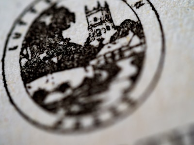 A close-up view of a circular black and white stamp featuring a medieval-style building, possibly a castle, with trees and a house in the background. The details are slightly blurred, emphasizing the texture of the paper.