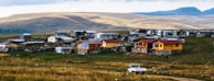 A rural settlement situated on a gently sloping hillside features a collection of small, modest houses made of wood and metal, some with vibrant yellow or red accents. A dirt road runs through the area, and a white vehicle is parked nearby. The landscape is expansive, with open grasslands and distant hills under a cloudy sky.