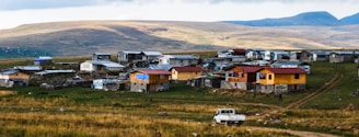 A rural settlement situated on a gently sloping hillside features a collection of small, modest houses made of wood and metal, some with vibrant yellow or red accents. A dirt road runs through the area, and a white vehicle is parked nearby. The landscape is expansive, with open grasslands and distant hills under a cloudy sky.