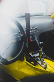 A car interior featuring a racing-style steering wheel and a prominent handbrake lever. The dashboard displays various control switches, and the cabin has a sleek, sporty appearance with visible yellow sections.