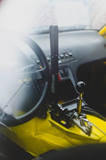 A car interior featuring a racing-style steering wheel and a prominent handbrake lever. The dashboard displays various control switches, and the cabin has a sleek, sporty appearance with visible yellow sections.