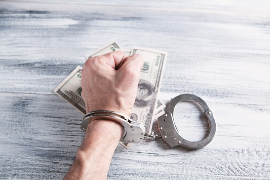 A hand wearing handcuffs is positioned on top of a pile of US dollar bills on a textured wooden surface. The handcuffs are open, suggesting recent use or potential freedom, and the hand is clenched into a fist.
