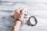 A hand wearing handcuffs is positioned on top of a pile of US dollar bills on a textured wooden surface. The handcuffs are open, suggesting recent use or potential freedom, and the hand is clenched into a fist.