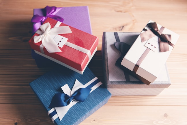 Several elegantly wrapped gift boxes are arranged on a wooden surface. They feature ribbons and bows in various colors, including purple, white, blue, and brown.