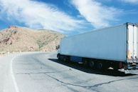 A large white semi-truck is driving along a curved mountain road under a clear blue sky. The landscape features arid, rocky hills with sparse vegetation. The road is clean and well-paved, with traffic signs visible, indicating a turn.