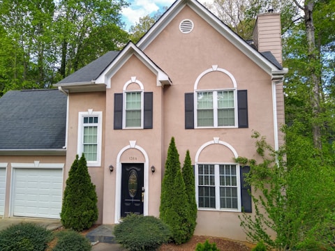A two-story house with a beige facade and black shutters on the windows. The front door is black and has a decorative glass panel. A garage with a white door is attached to the left side of the house. The front yard is landscaped with shrubs and small trees, creating a cozy and well-maintained appearance. The house is surrounded by tall, green trees and a cloudy sky can be seen in the background.