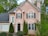 A two-story house with a beige facade and black shutters on the windows. The front door is black and has a decorative glass panel. A garage with a white door is attached to the left side of the house. The front yard is landscaped with shrubs and small trees, creating a cozy and well-maintained appearance. The house is surrounded by tall, green trees and a cloudy sky can be seen in the background.