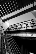 An architectural photograph featuring a building captured from a low angle, emphasizing its geometric lines and modern design. The image is in black and white, highlighting contrasts and shadows. The composition includes structural elements like metal railings and beams intersecting in dynamic angles.