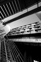 An architectural photograph featuring a building captured from a low angle, emphasizing its geometric lines and modern design. The image is in black and white, highlighting contrasts and shadows. The composition includes structural elements like metal railings and beams intersecting in dynamic angles.