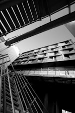 An architectural photograph featuring a building captured from a low angle, emphasizing its geometric lines and modern design. The image is in black and white, highlighting contrasts and shadows. The composition includes structural elements like metal railings and beams intersecting in dynamic angles.