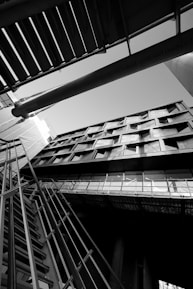 An architectural photograph featuring a building captured from a low angle, emphasizing its geometric lines and modern design. The image is in black and white, highlighting contrasts and shadows. The composition includes structural elements like metal railings and beams intersecting in dynamic angles.