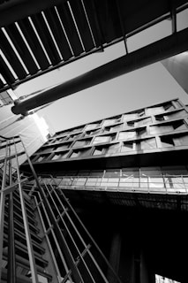An architectural photograph featuring a building captured from a low angle, emphasizing its geometric lines and modern design. The image is in black and white, highlighting contrasts and shadows. The composition includes structural elements like metal railings and beams intersecting in dynamic angles.