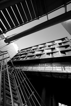 An architectural photograph featuring a building captured from a low angle, emphasizing its geometric lines and modern design. The image is in black and white, highlighting contrasts and shadows. The composition includes structural elements like metal railings and beams intersecting in dynamic angles.