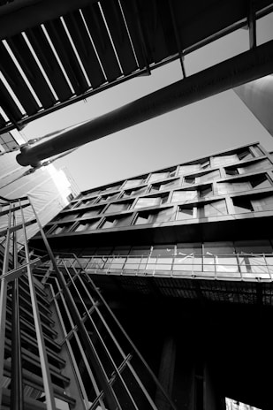 An architectural photograph featuring a building captured from a low angle, emphasizing its geometric lines and modern design. The image is in black and white, highlighting contrasts and shadows. The composition includes structural elements like metal railings and beams intersecting in dynamic angles.