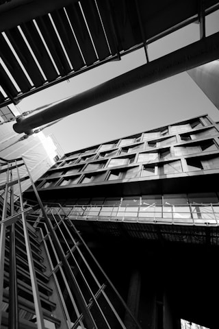 An architectural photograph featuring a building captured from a low angle, emphasizing its geometric lines and modern design. The image is in black and white, highlighting contrasts and shadows. The composition includes structural elements like metal railings and beams intersecting in dynamic angles.