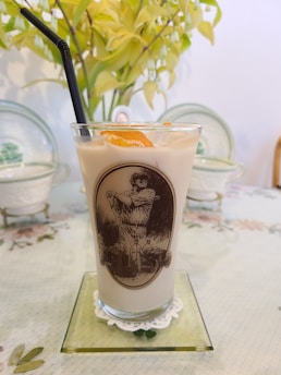 A glass of iced milk tea with a black straw, adorned with an illustration featuring a historical figure and some men in hats. An orange slice is placed on top of the drink. The glass sits on a glass coaster with a doily underneath, on a table covered with a floral tablecloth. In the background, there are two decorative plates and a plant with yellowish-green leaves.