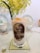 A glass of iced milk tea with a black straw, adorned with an illustration featuring a historical figure and some men in hats. An orange slice is placed on top of the drink. The glass sits on a glass coaster with a doily underneath, on a table covered with a floral tablecloth. In the background, there are two decorative plates and a plant with yellowish-green leaves.