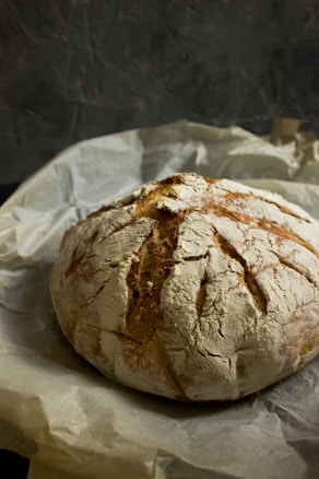A round loaf of rustic bread with a textured, cracked crust sits on a piece of parchment paper. The bread has a golden-brown finish with flour dusting, and its surface shows depth and character through several natural cracks.