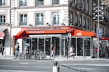 A bustling street corner features a café with a bright orange awning labeled 'café Madeleine.' Several people are walking by, some with bicycles parked in front of the building. The architecture of the surrounding buildings is classic and European, with large windows and ornate balconies. The scene is lively, with a sense of urban activity.
