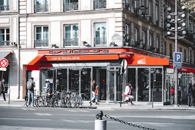 A bustling street corner features a café with a bright orange awning labeled 'café Madeleine.' Several people are walking by, some with bicycles parked in front of the building. The architecture of the surrounding buildings is classic and European, with large windows and ornate balconies. The scene is lively, with a sense of urban activity.