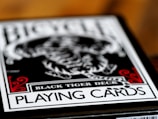 A close-up of a playing card box labeled Black Tiger Deck, featuring bold text and intricate design elements with a focus on a monochrome tiger illustration. Red decorative accents frame the corners while the overall color scheme is black and white.