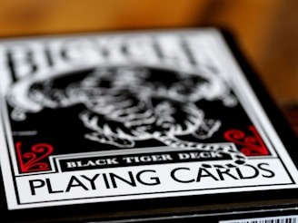 A close-up of a playing card box labeled Black Tiger Deck, featuring bold text and intricate design elements with a focus on a monochrome tiger illustration. Red decorative accents frame the corners while the overall color scheme is black and white.