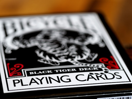 A close-up of a playing card box labeled Black Tiger Deck, featuring bold text and intricate design elements with a focus on a monochrome tiger illustration. Red decorative accents frame the corners while the overall color scheme is black and white.