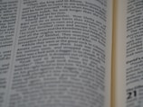 A close-up view of an open book with text focusing on biblical scripture. The text is printed in two columns on a thin, slightly yellowed page, indicating an older or traditional style publication. The lighting highlights parts of the text while leaving other areas slightly shadowed.