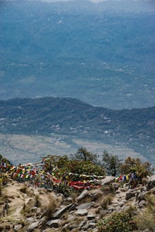 A scenic view of a mountainous landscape with lush greenery and colorful prayer flags hanging across rocky terrain. In the distance, rolling hills and a vast valley with scattered houses are visible, under a slightly overcast sky.