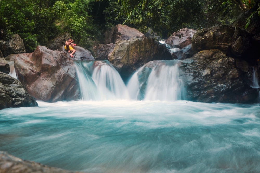 man in yellow jacket sitting on rock near waterfalls during daytime