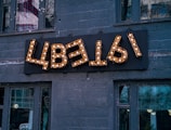 A decorative sign with Cyrillic letters mounted on a dark gray brick wall, illuminated by small light bulbs. The building has several large windows, reflecting urban surroundings and trees.