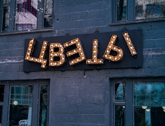 A decorative sign with Cyrillic letters mounted on a dark gray brick wall, illuminated by small light bulbs. The building has several large windows, reflecting urban surroundings and trees.