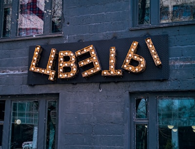 A decorative sign with Cyrillic letters mounted on a dark gray brick wall, illuminated by small light bulbs. The building has several large windows, reflecting urban surroundings and trees.