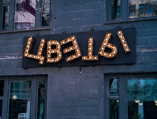 A decorative sign with Cyrillic letters mounted on a dark gray brick wall, illuminated by small light bulbs. The building has several large windows, reflecting urban surroundings and trees.