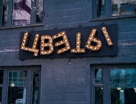 A decorative sign with Cyrillic letters mounted on a dark gray brick wall, illuminated by small light bulbs. The building has several large windows, reflecting urban surroundings and trees.