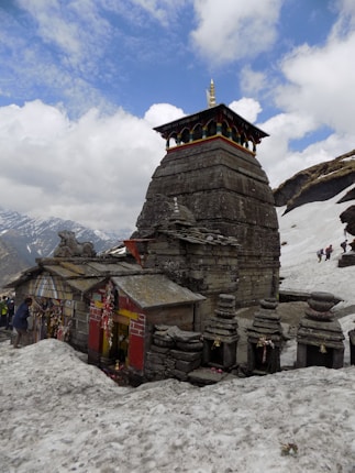 A stone temple structure with traditional architecture stands amidst a snowy landscape. The temple has intricate carvings and is adorned with colorful elements, including a small spire on top. Surrounding stones are arranged carefully, and a few small bells are attached to the structure. People can be seen in the distance, climbing or walking in the snow-covered hills.