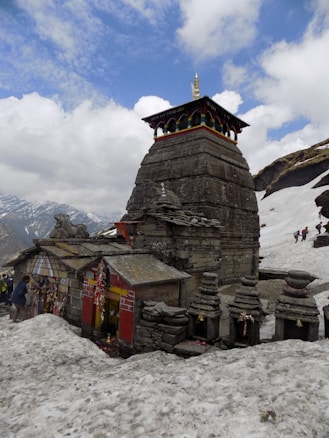 A stone temple structure with traditional architecture stands amidst a snowy landscape. The temple has intricate carvings and is adorned with colorful elements, including a small spire on top. Surrounding stones are arranged carefully, and a few small bells are attached to the structure. People can be seen in the distance, climbing or walking in the snow-covered hills.