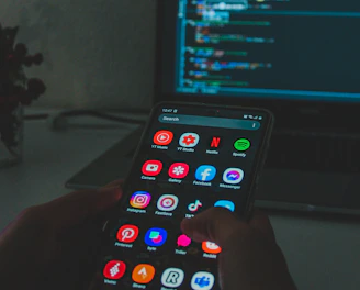 A person holds a smartphone displaying various colorful app icons, placed in front of a laptop screen showing a code editor with lines of code. The setting appears dimly lit, suggesting a focus on technology and coding.